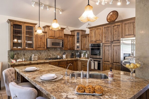 Una cocina con gabinetes de madera, encimeras de granito, electrodomésticos de acero inoxidable e iluminación colgante.