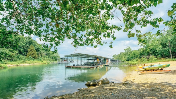 A riverside dock with a covered structure, calm water, and a gravel path leading to a boat. - Tribesman Resort #9 - Renovated Condo, Lake Access, Slip Rental, Great Location! (Branson)