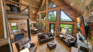 A spacious loft-style living area with a high ceiling, large windows, and a cozy fireplace. - Mountain Cabin - Views, Theater, Game Room, Pool (Sevierville)