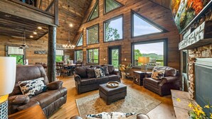 A spacious living room with leather sofas, a fireplace, and large windows offering a view of the outdoors. - Mountain Cabin - Views, Theater, Game Room, Pool (Sevierville)