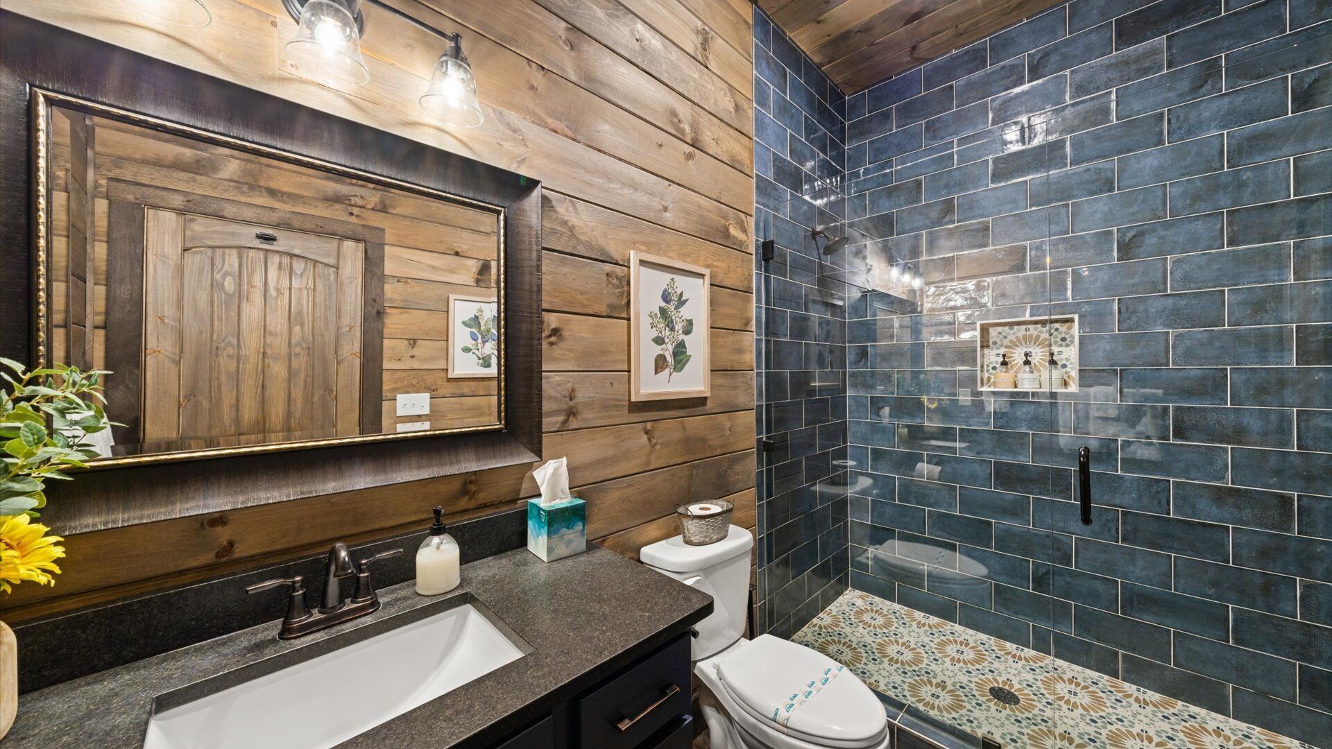 A bathroom with a wooden vanity, a sink, a toilet, and a shower area with dark tiles.