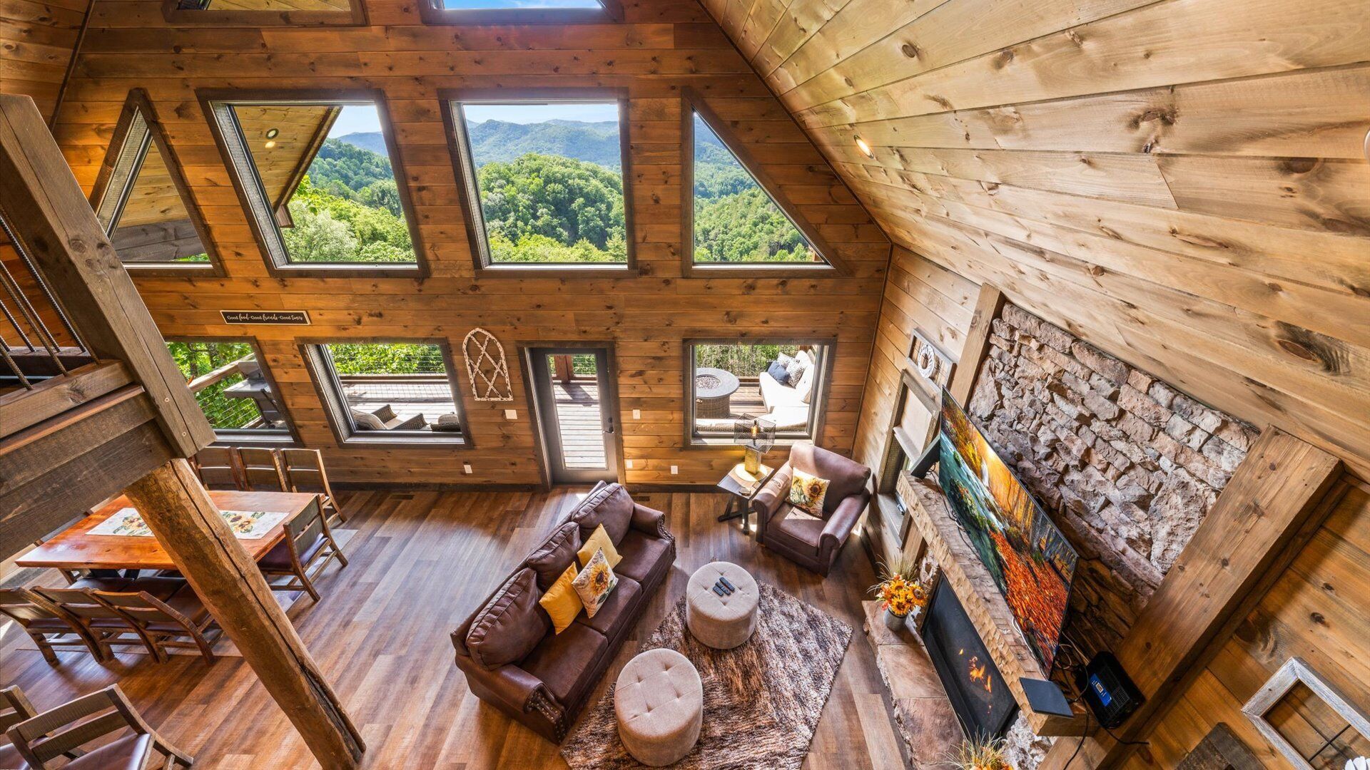 Amplio espacio tipo loft con techos altos, grandes ventanales que ofrecen vistas a un paisaje boscoso, una zona de estar acogedora con chimenea y una pared de piedra con obras de arte.
