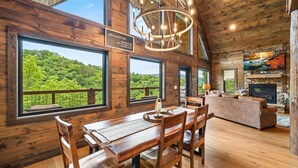 Interior rústico de cabana com paredes de madeira, janelas amplas com vista para a floresta, área de jantar, sala de estar com lareira e um lustre.