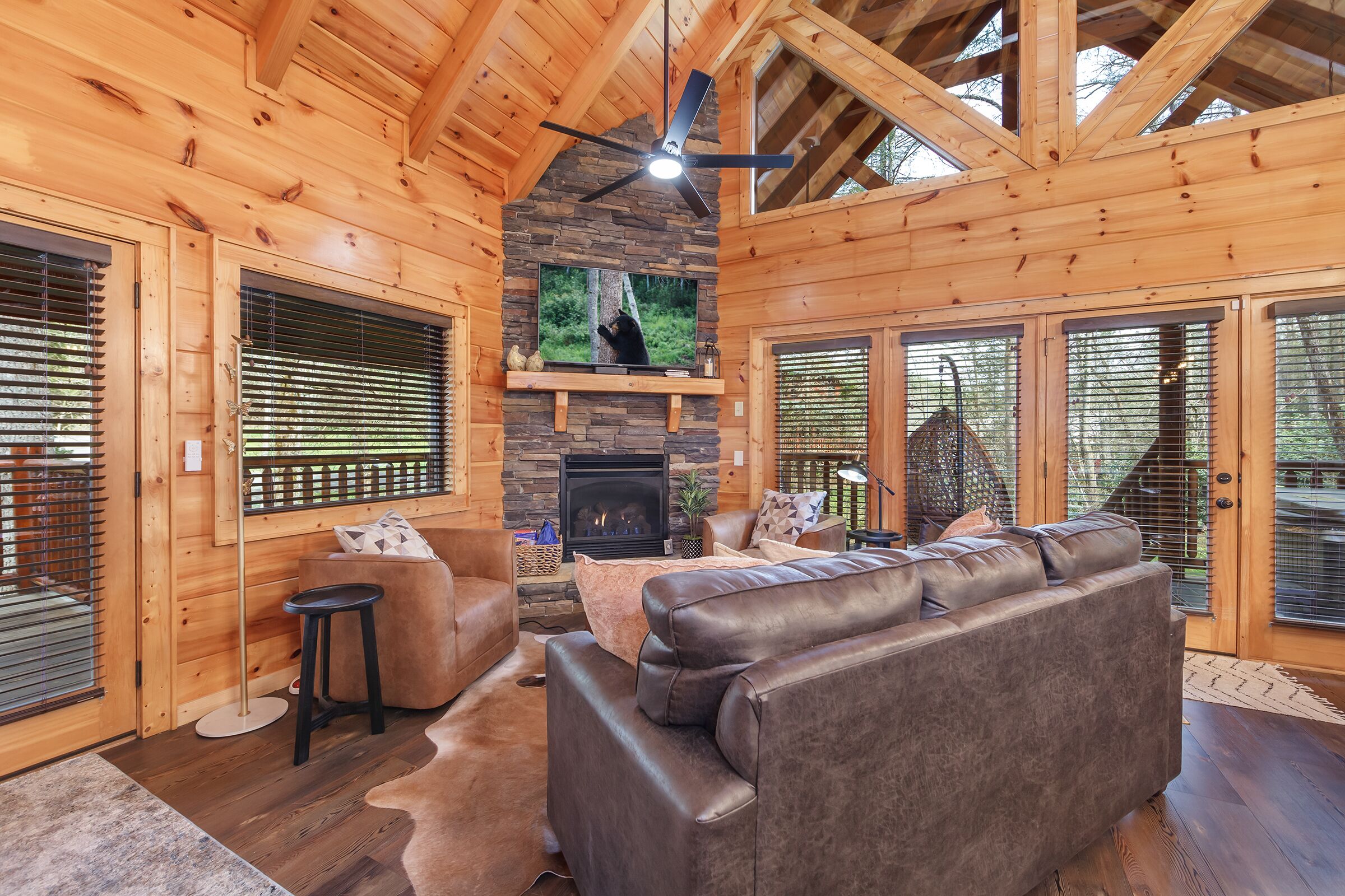 A cozy log cabin interior with a stone fireplace, leather sofa, and a large TV screen.