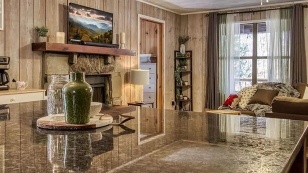 A spacious living area with a wooden interior, a fireplace, a large window with curtains, and a cozy seating area. - *Romantic 3BD w/ HOT TUB! Nestled in the WOODS!* (Pigeon Forge)