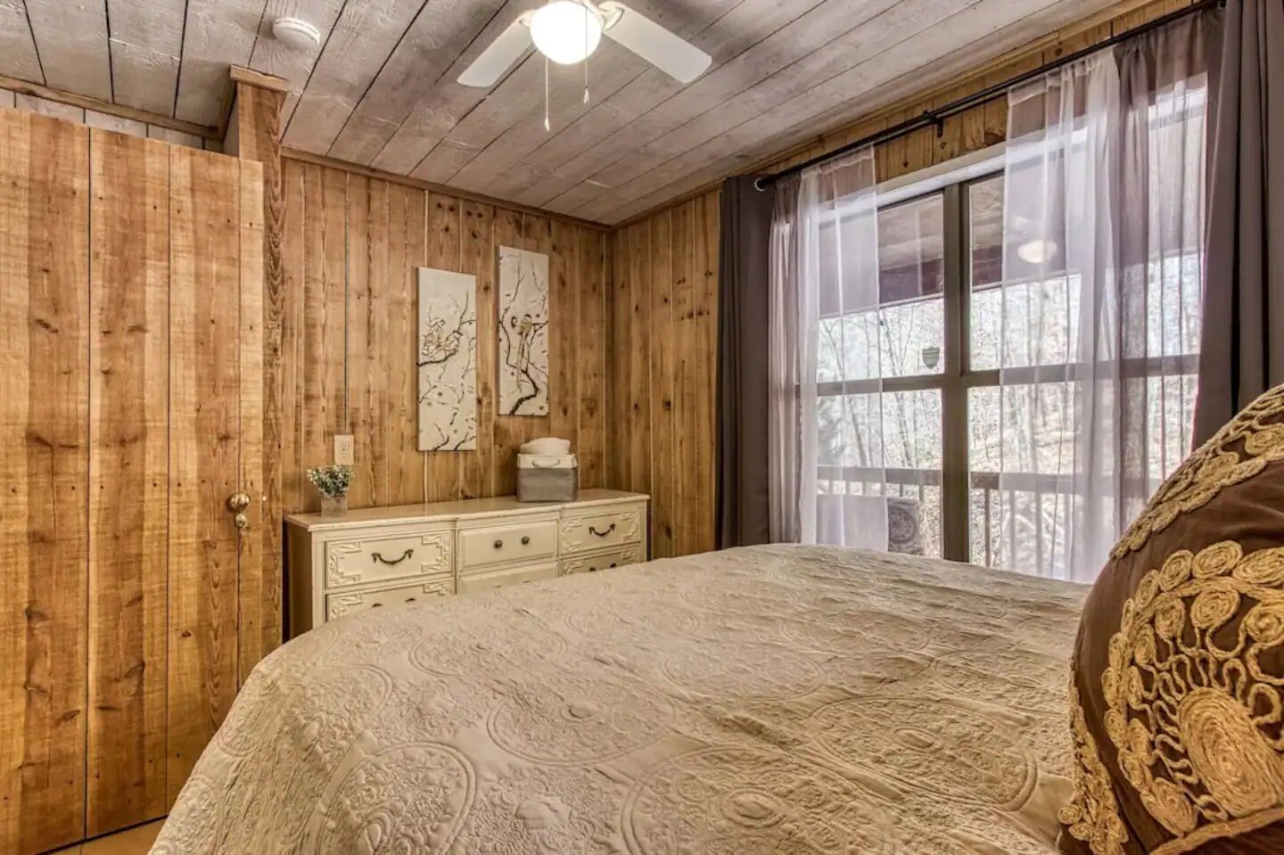 Quarto com paredes de madeira, cama com colcha estampada, cômoda e janela com cortinas.