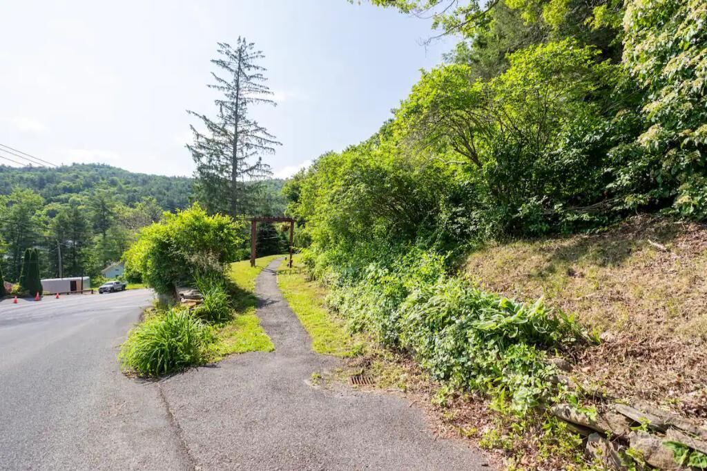 A paved path alongside a road with dense greenery and a wooden structure on the right.