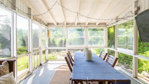 Une terrasse couverte avec une longue table et des chaises, de grandes fenêtres et des guirlandes lumineuses.