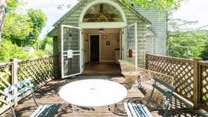 Une terrasse en bois avec une table et des chaises blanches, un patio couvert et une vue sur une maison dont la porte est ouverte.