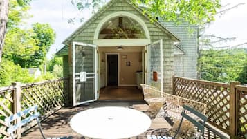 Une terrasse en bois avec une table et des chaises blanches, un patio couvert et une vue sur une maison dont la porte est ouverte.