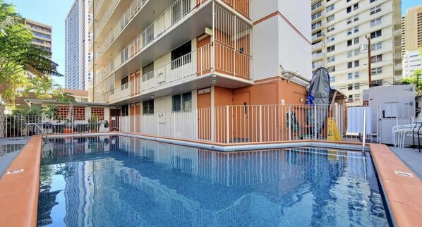 Una piscina con borde de baldosas, rodeada por una valla blanca y un edificio con balcones.