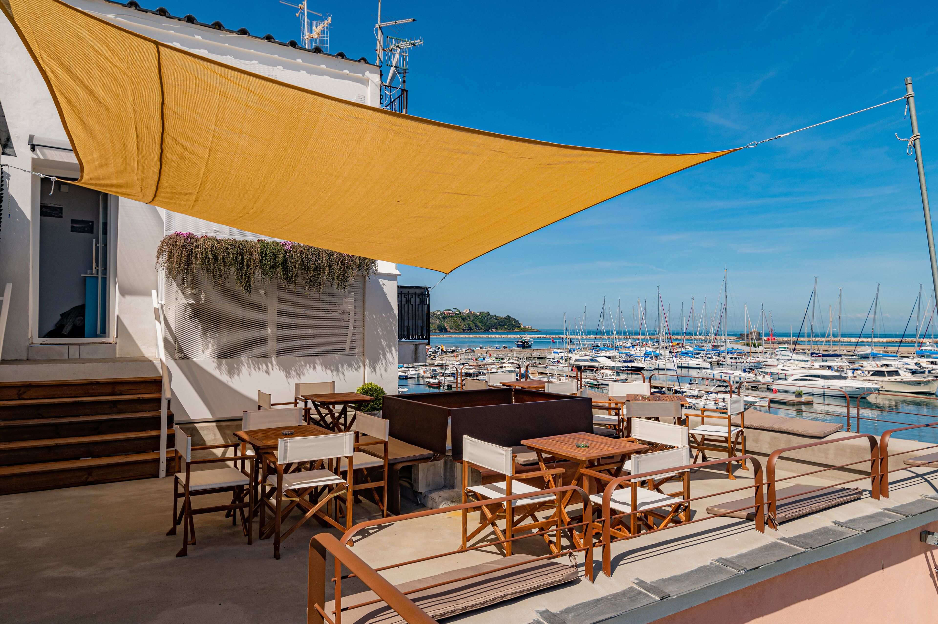 An outdoor seating area with wooden furniture and a yellow shade sail, overlooking a marina with numerous boats.