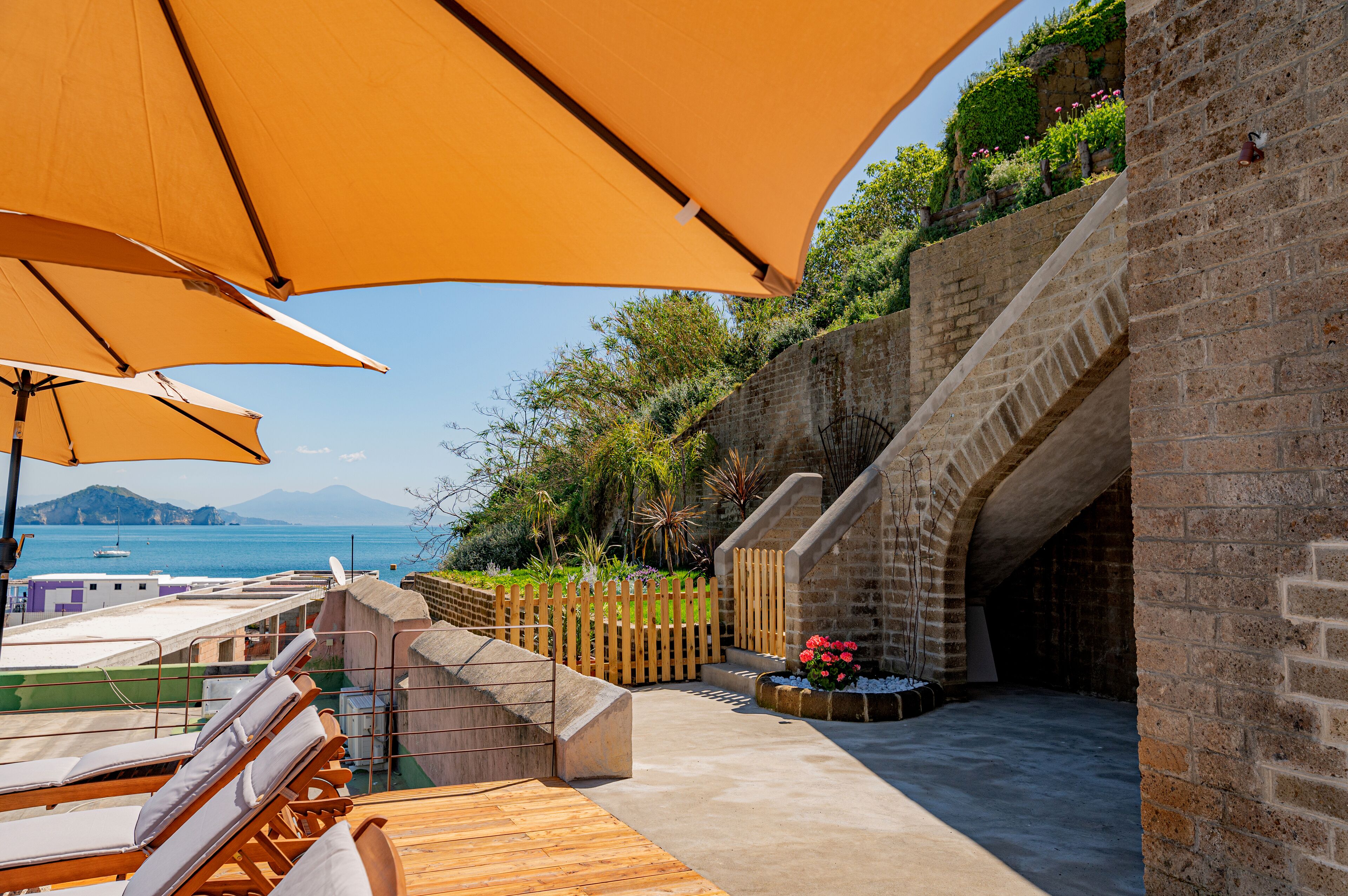 A terrace with wooden lounge chairs and large umbrellas, a stone wall with a staircase, and a view of the sea and mountains.