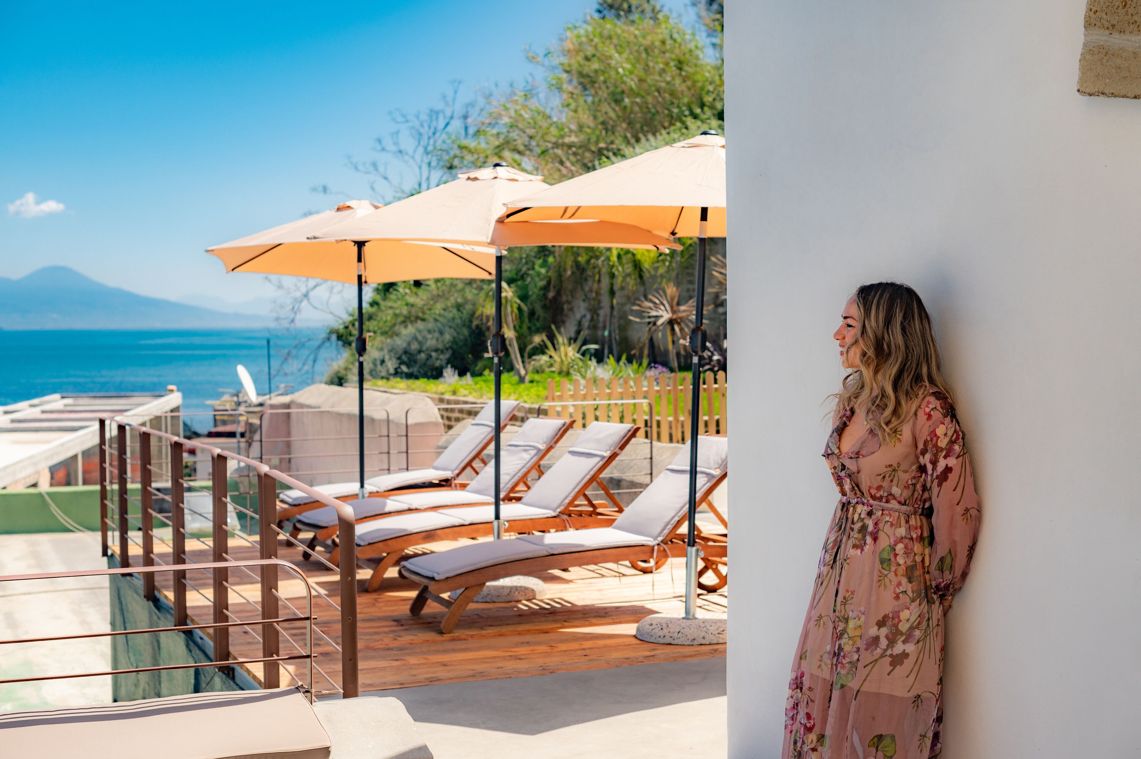 Un área junto a la piscina con sillones y sombrillas, una terraza de madera y vista al mar.