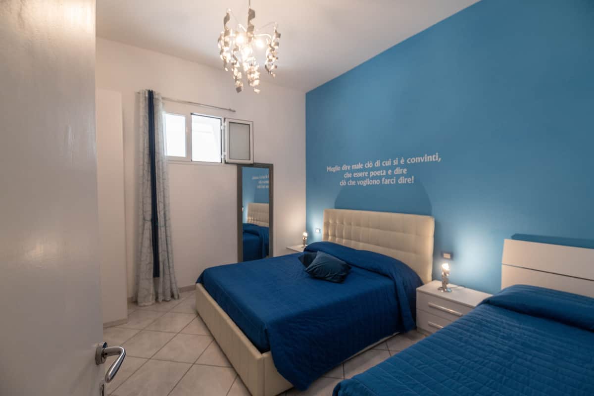 A bedroom with a blue accent wall, a chandelier, a bed with blue bedding, and a mirror on the wall.