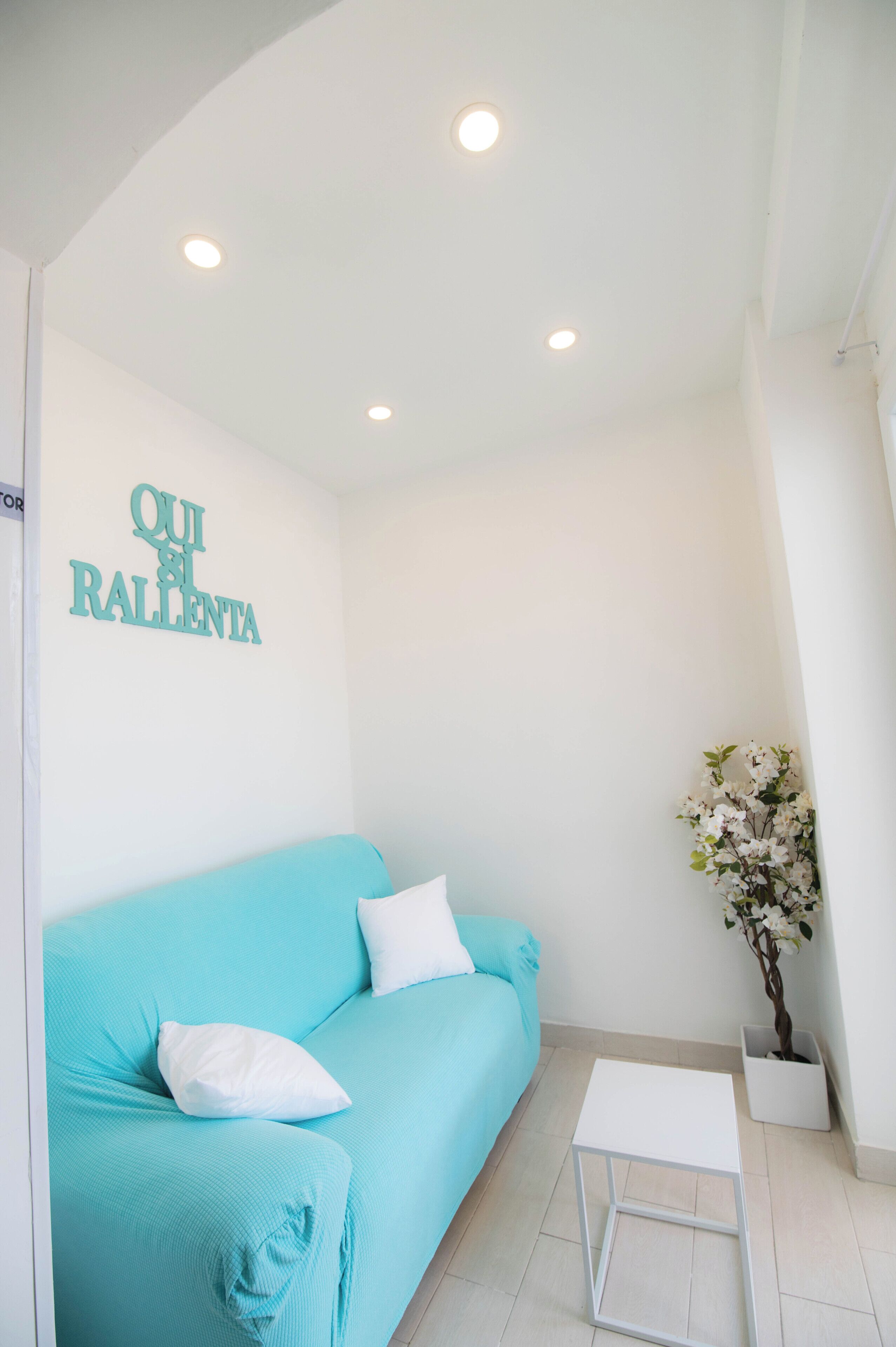 A room with a turquoise sofa, a small white table, a potted plant, and a wall with the text 'QUI ES RALLENTA'.