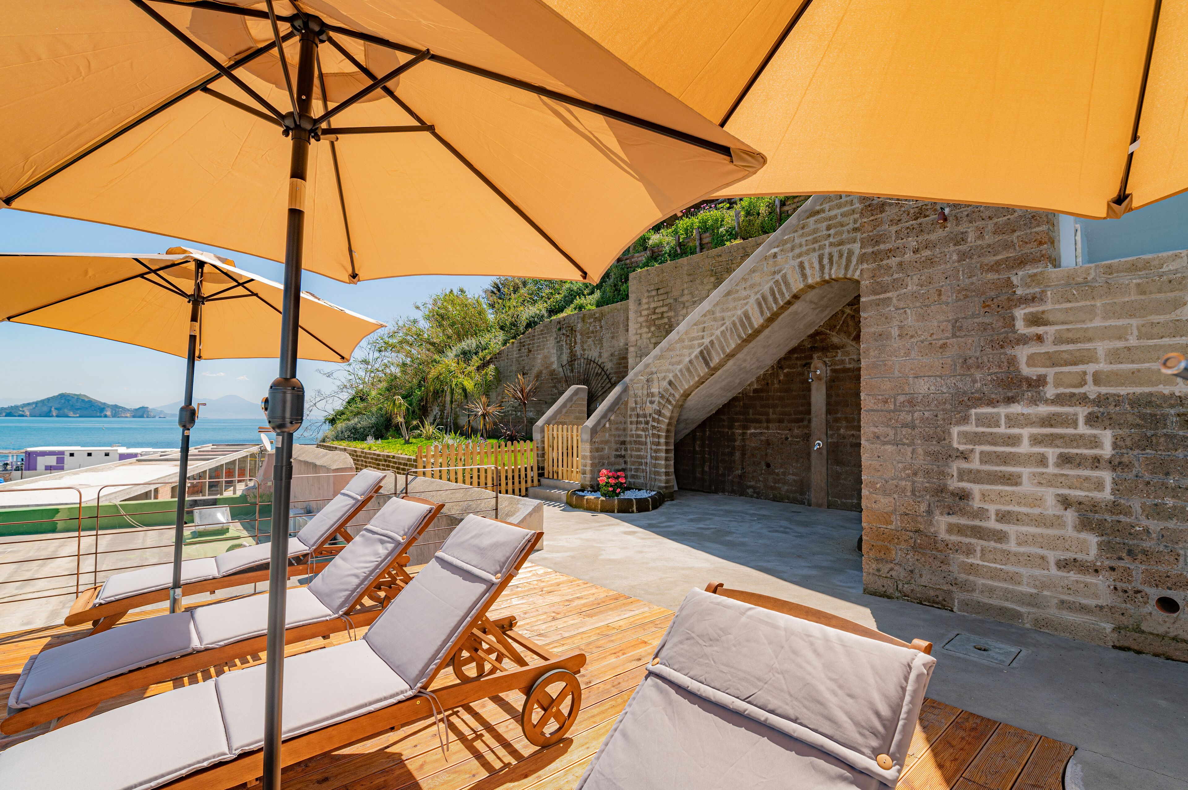 A sunlit outdoor seating area with wooden loungers and large umbrellas, overlooking a pool and a scenic view of the sea.