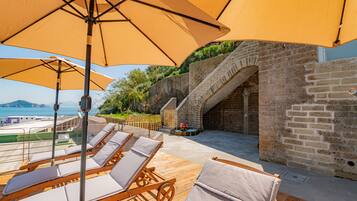 A sunlit outdoor seating area with wooden loungers and large umbrellas, overlooking a pool and a scenic view of the sea.