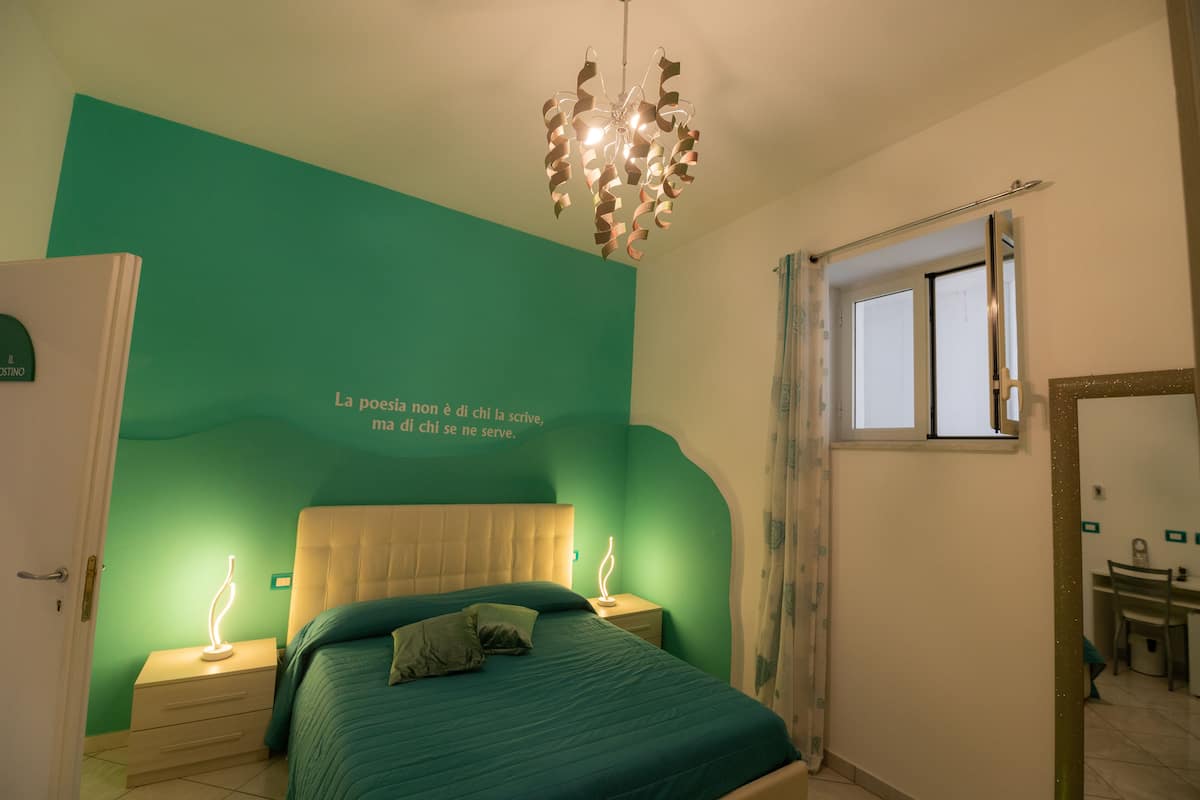 A bedroom with teal walls, a bed with a white headboard, two bedside tables with lamps, a window with curtains, and a mirror on the wall.