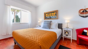Une chambre d’hôtel avec un grand lit, deux tables de chevet, un fauteuil rouge, une œuvre d’art accrochée au mur et une fenêtre avec des rideaux.