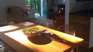 En matplats med ett träbord, stolar och en skål med frukt. I bakgrunden står en tv på ett stativ och en person sitter och använder en bärbar dator.