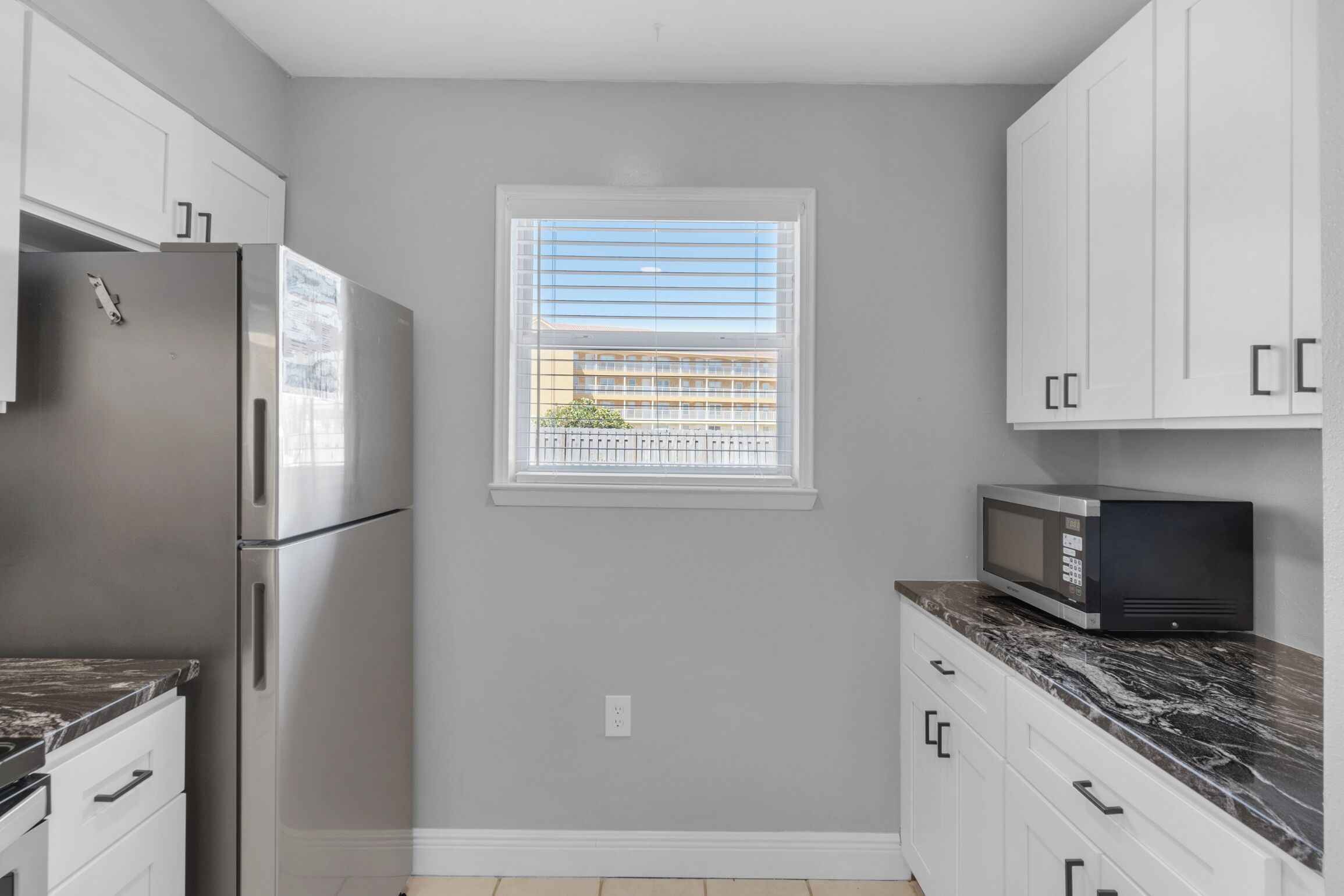 A compact kitchen with white cabinets, a stainless steel refrigerator, a microwave, and a window with blinds.