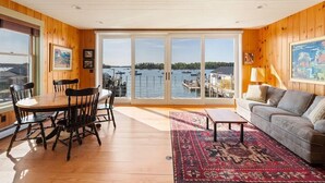 A spacious room with a wooden interior, a dining table with chairs, a sofa, and a large window offering a view of boats and water.