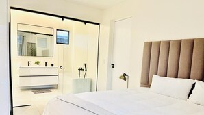 Une chambre moderne avec un grand lit, une table de chevet, un miroir et une salle de bain équipée d’un lavabo et d’une douche.