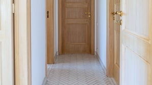 Un couloir se terminant par une porte en bois, flanqué de portes en bois de chaque côté, et dont le sol est recouvert d’un parquet à motifs en chevrons.