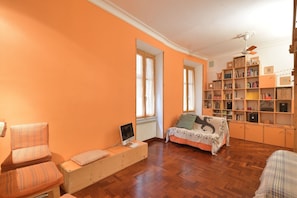 Une pièce avec un mur orange, un plancher en bois, un lit avec une couverture, une chaise, un bureau avec un ordinateur et une bibliothèque.