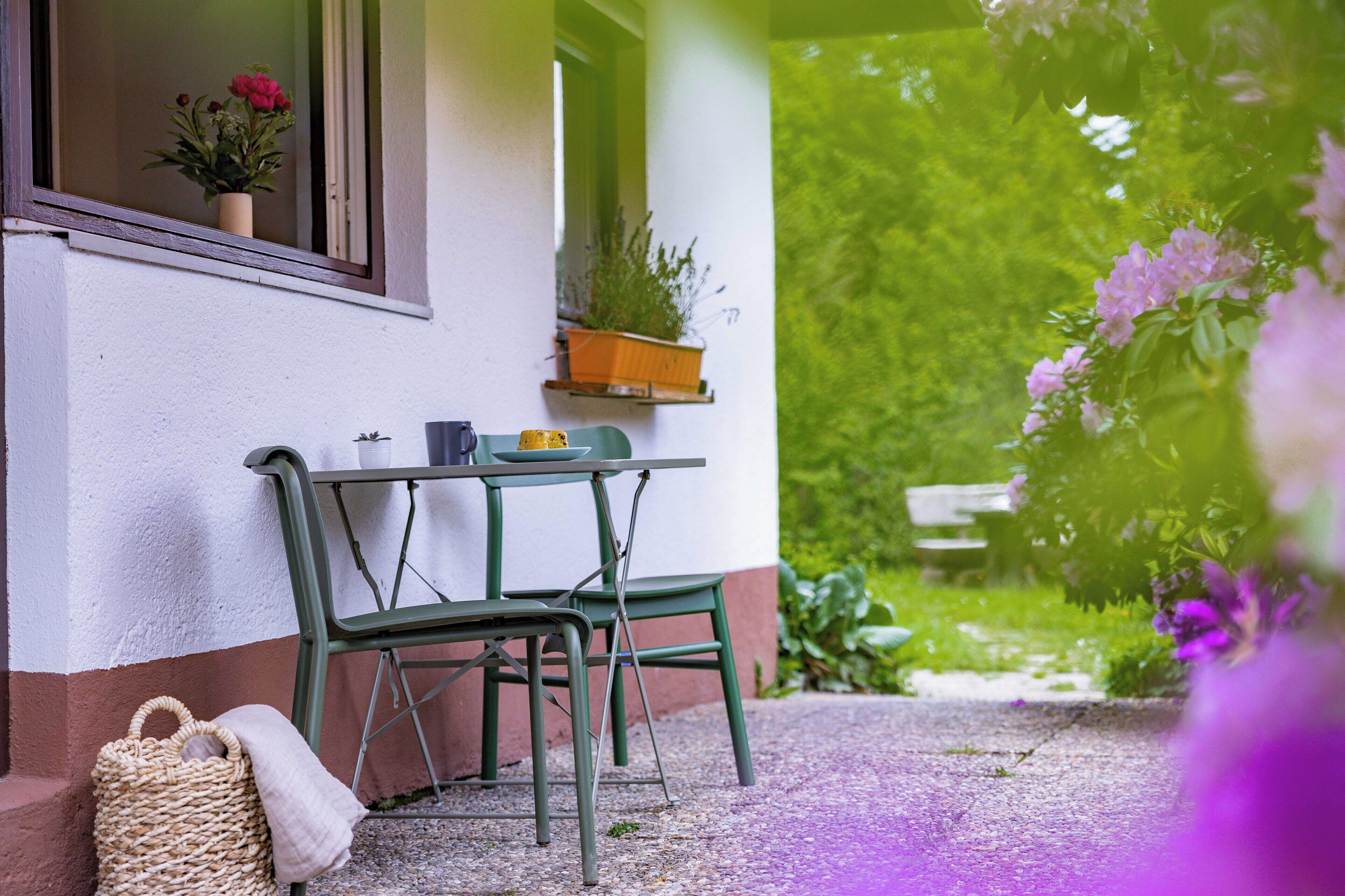 Une table et des chaises en métal vert se trouvent à l’extérieur d’un bâtiment. Une fenêtre donne sur une plante en pot.
