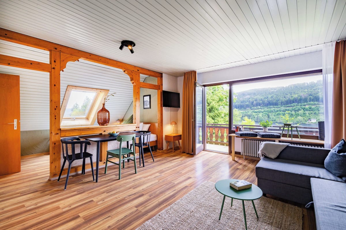 A cozy room with a wooden ceiling, a sofa, a dining table with chairs, and a large window offering a view of a forested area.