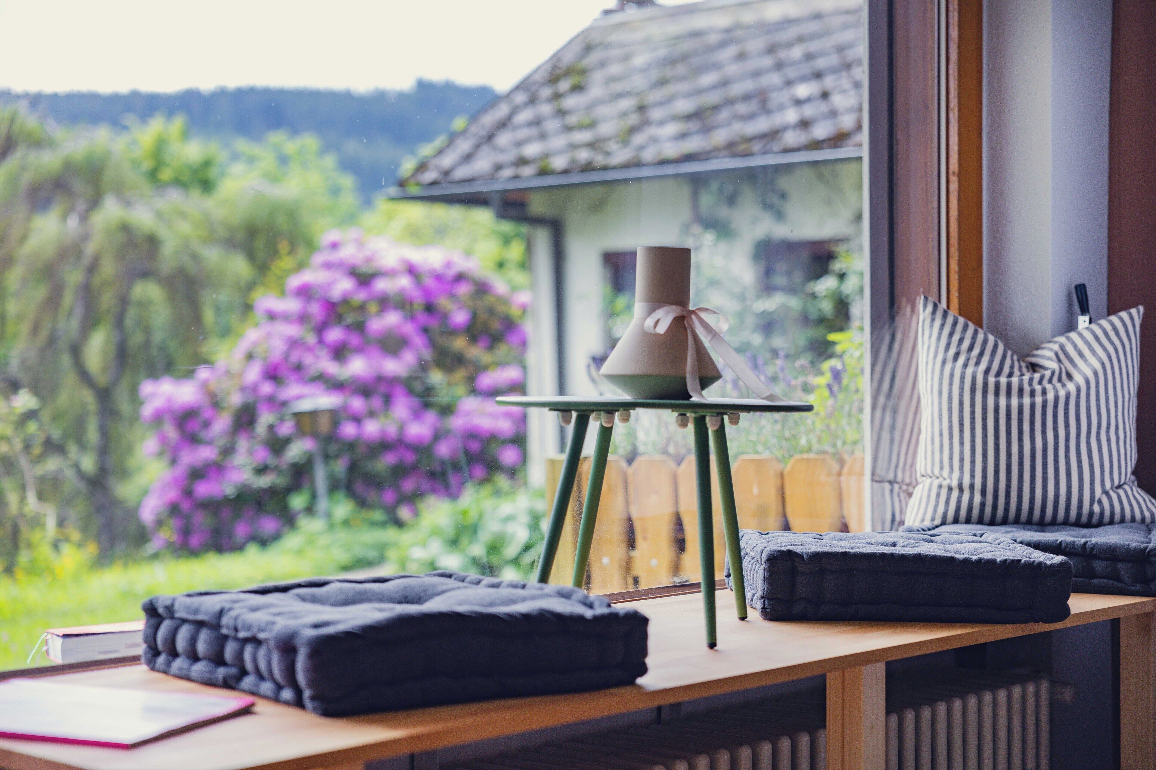 Une pièce chaleureuse avec une table en bois, une chaise, une petite table sur laquelle est posée une bougie, et une vue sur une maison et un jardin.