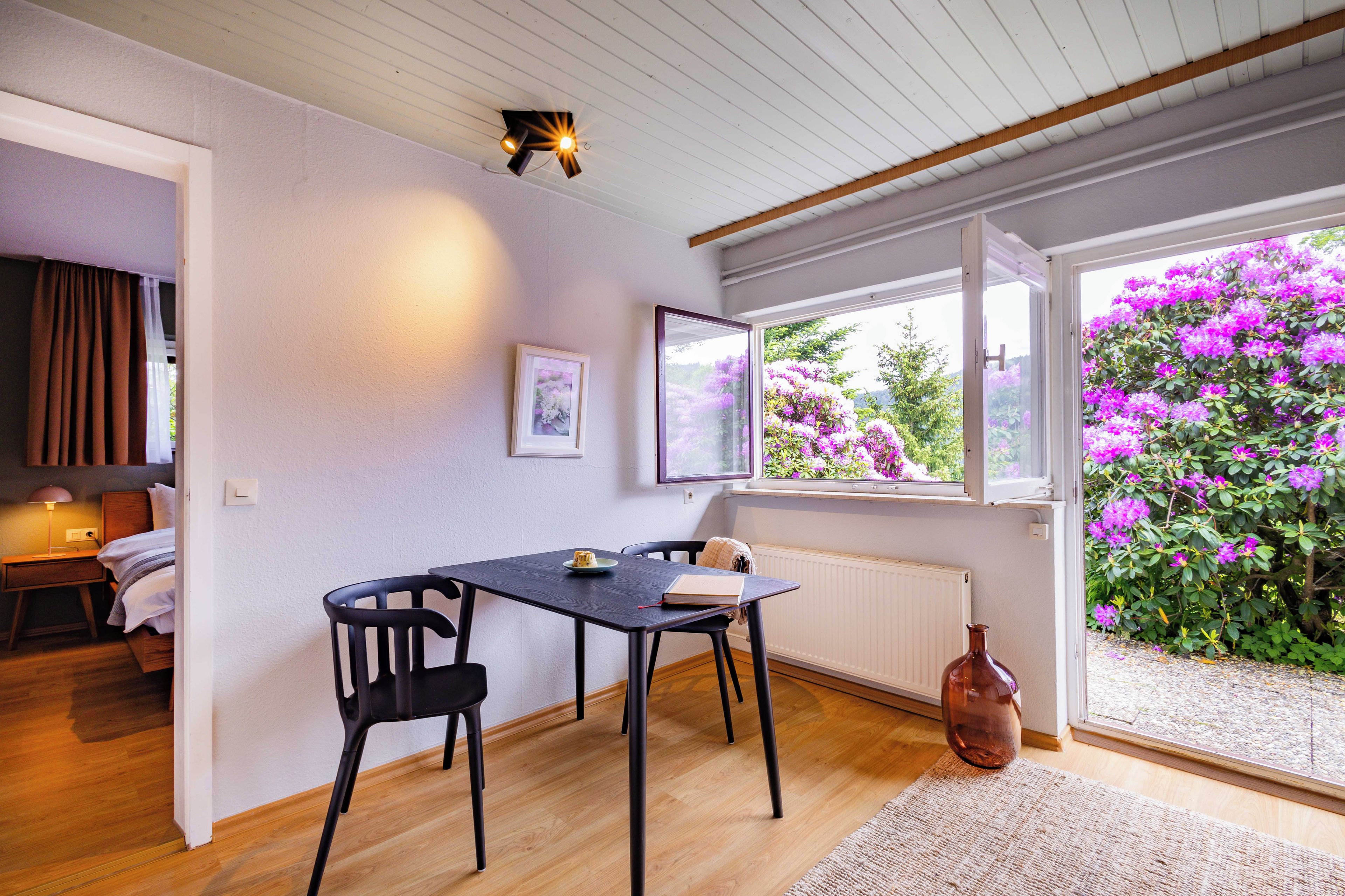 Une pièce comprenant une table à manger, des chaises, un lit, une fenêtre donnant sur un parterre de fleurs violettes et un luminaire au plafond.
