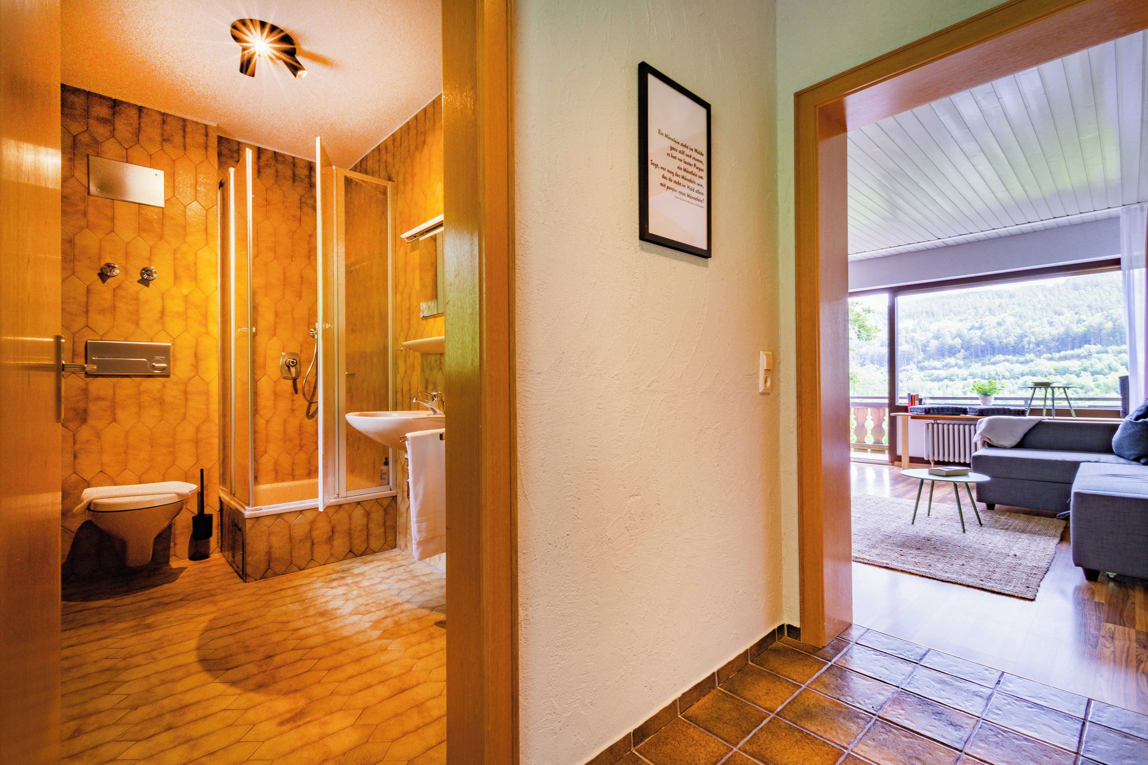 A modern bathroom with a shower, toilet, and sink. There is a framed quote on the wall and a view of a living area with a sofa and a table.