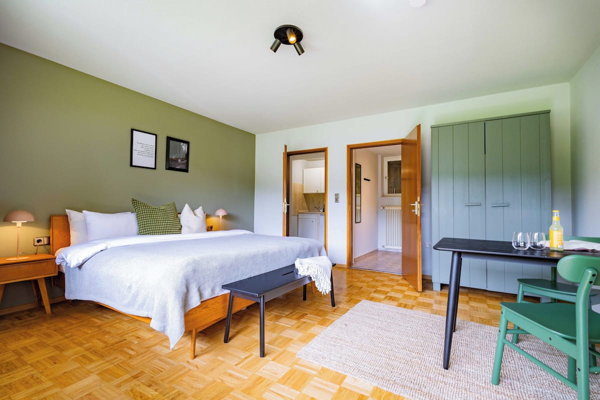 A bedroom with a bed, bedside table, and a desk with chairs. There is a door leading to a bathroom and a kitchen.