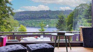 A room with a view of a lake and forest, a table with books and a glass, and two ottomans. - Titisee Haus an der Sonne by Kuckucksnester (Hinterzarten)