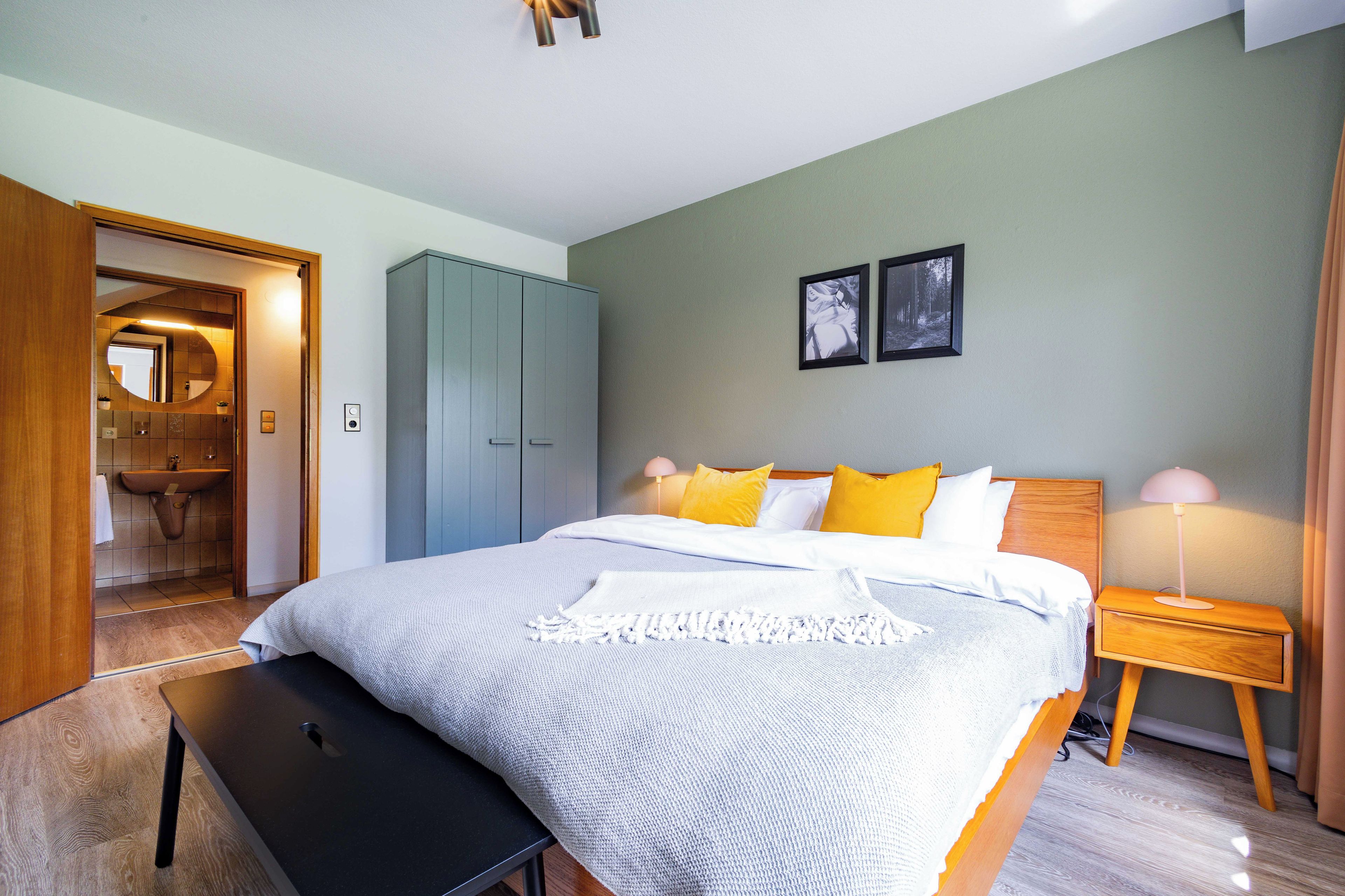 A bedroom with a bed, bedside tables, a bench, a wardrobe, and a bathroom visible through an open door.