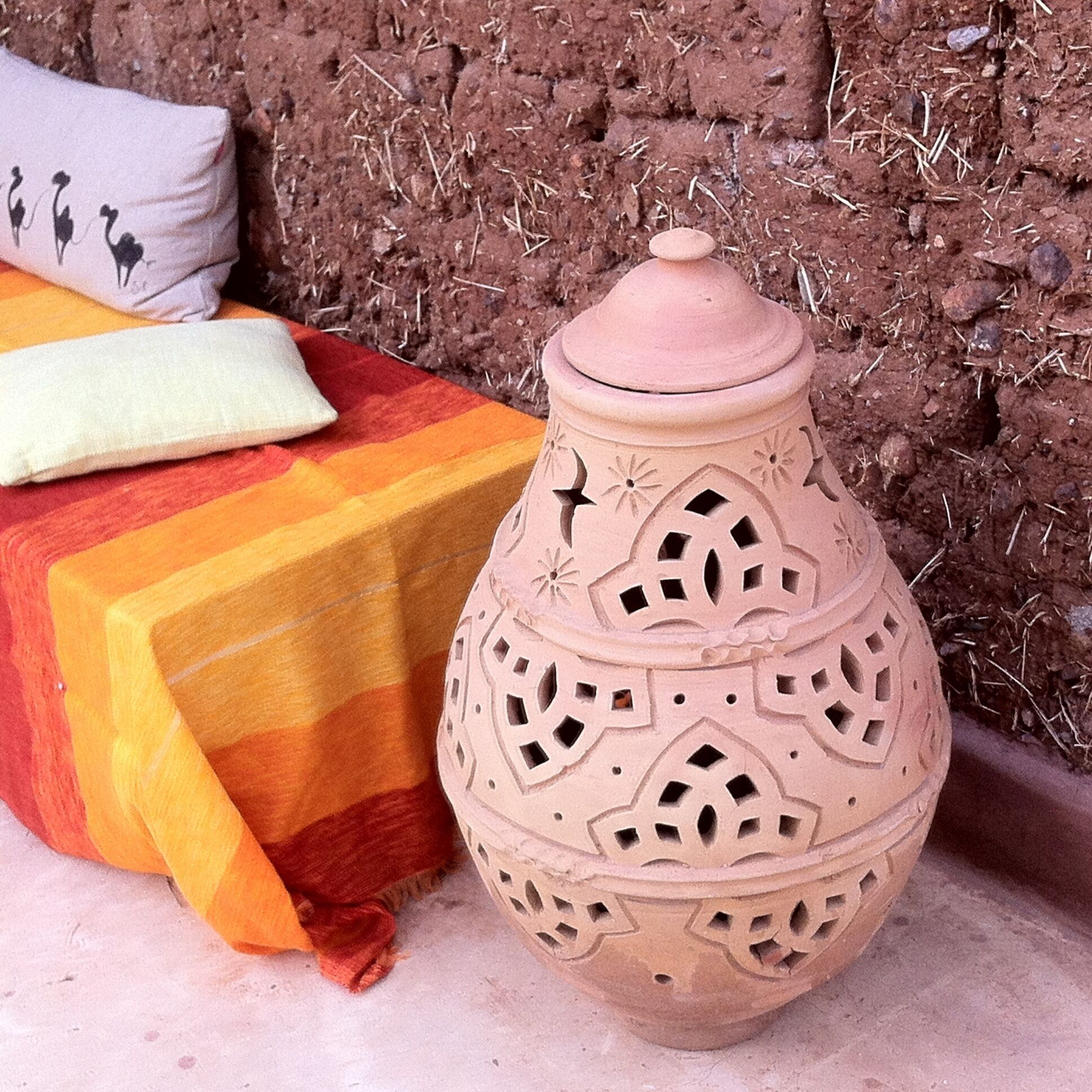 Un pot en terre cuite orné de motifs complexes découpés, posé sur une surface en béton, à côté d’un lit recouvert d’une couverture rayée aux couleurs vives et d’un coussin à motif d’oiseau.