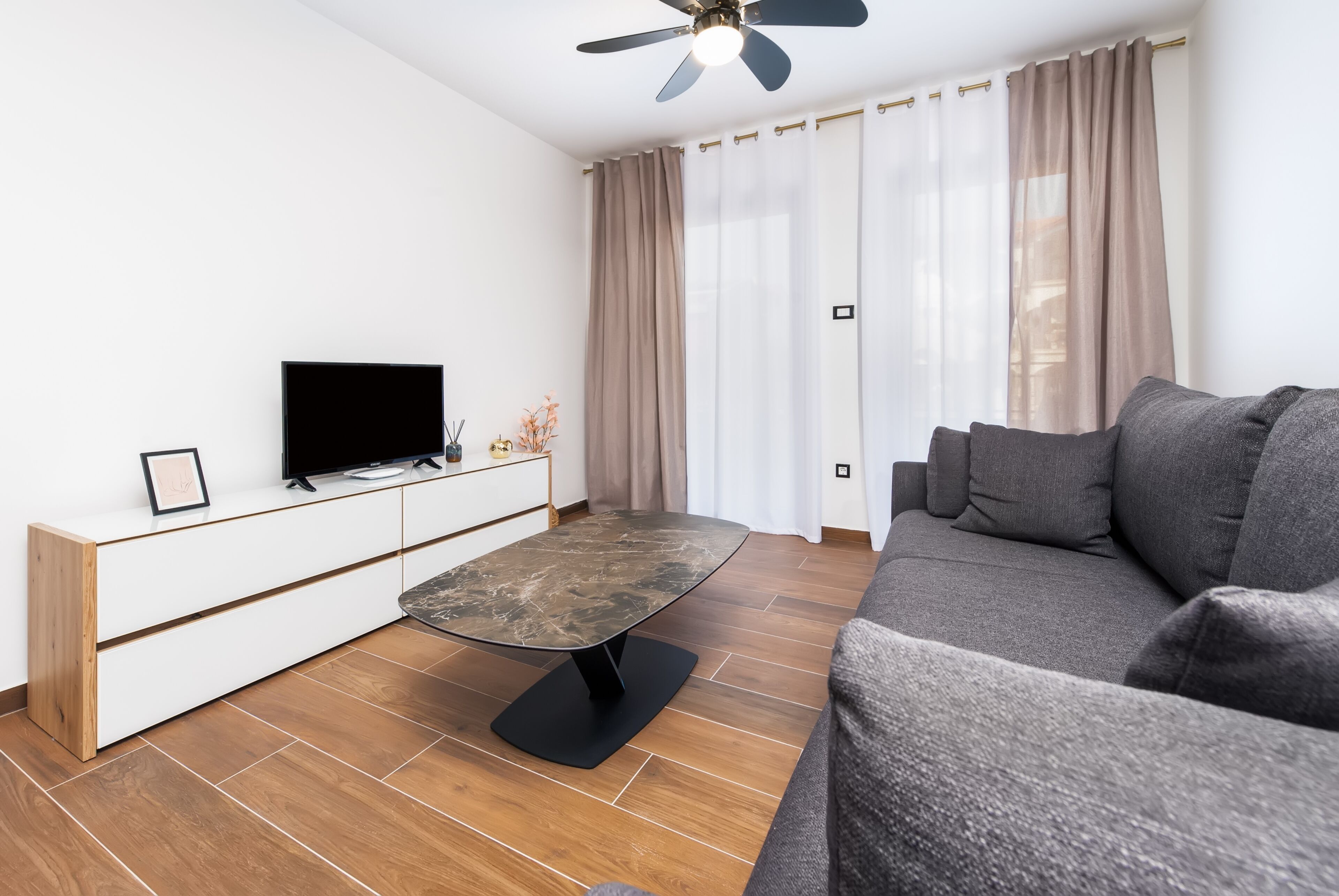 A modern living room with a flat-screen TV on a white console, a dark marble coffee table, a grey sofa, and a ceiling fan.