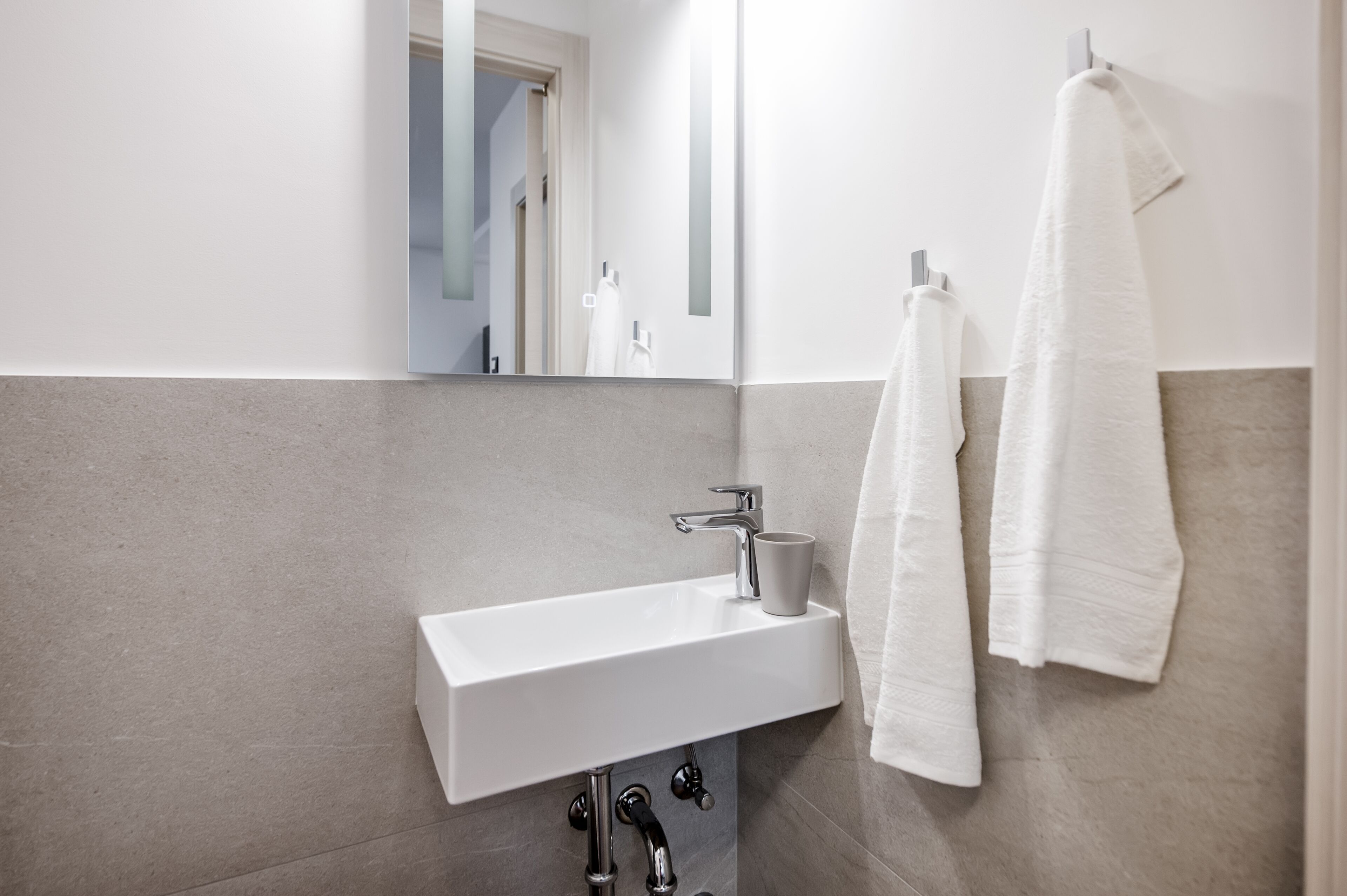 Un baño moderno con un lavamanos blanco, un espejo y dos toallas colgadas en un perchero.