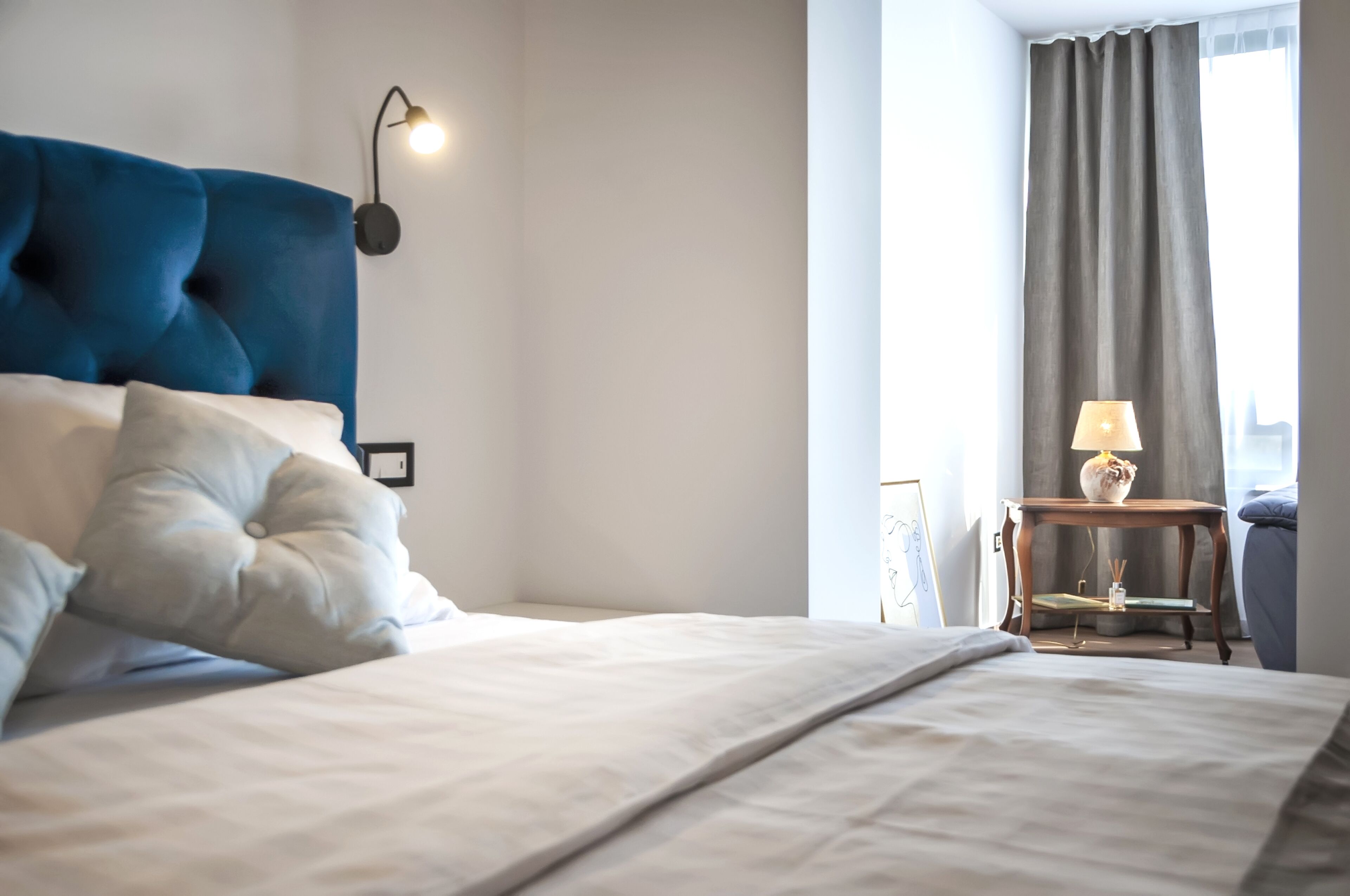 A neatly made bed with a blue headboard, a bedside table with a lamp, and a view of a window with curtains.