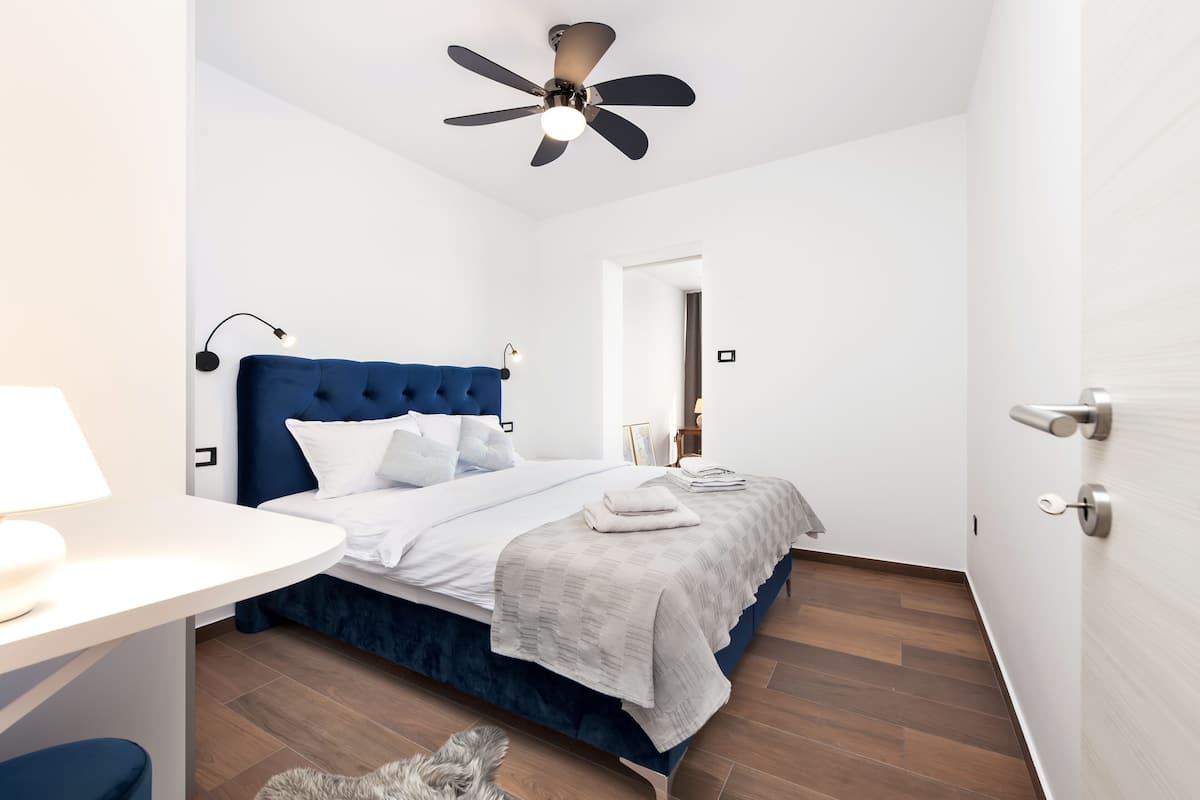 A modern bedroom with a blue upholstered headboard, a white bedspread, and a ceiling fan.