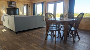 A cozy room with a grey sofa, wooden dining table with chairs, a TV, and large windows with curtains. - Peaceful quite cabin to kick back, relax and recharge! (Robert Lee)