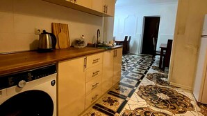 A kitchen with a washing machine, wooden cabinets, a tiled floor, and a dining area with a table and chairs. - Good Night (Tbilisi)
