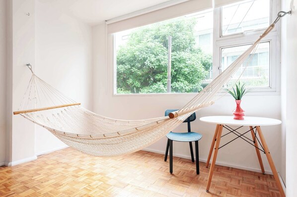 A hammock in a room with a wooden floor, a small round table with a potted plant, and a window with a view of greenery.