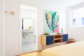 A modern bedroom with a bed, a wooden dresser, a colorful abstract painting, and a bathroom with a shower and a mirror.