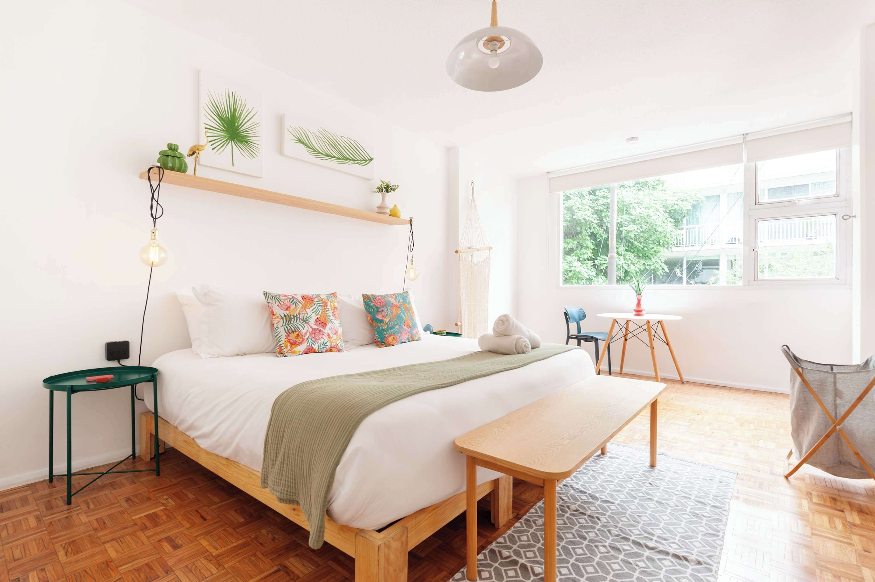 A modern bedroom with a wooden bed, a bedside table, a hanging light fixture, and a window with a view of greenery.