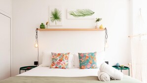 A bed with two pillows, a bedside table, a wall-mounted shelf with plants, and a hanging macrame.