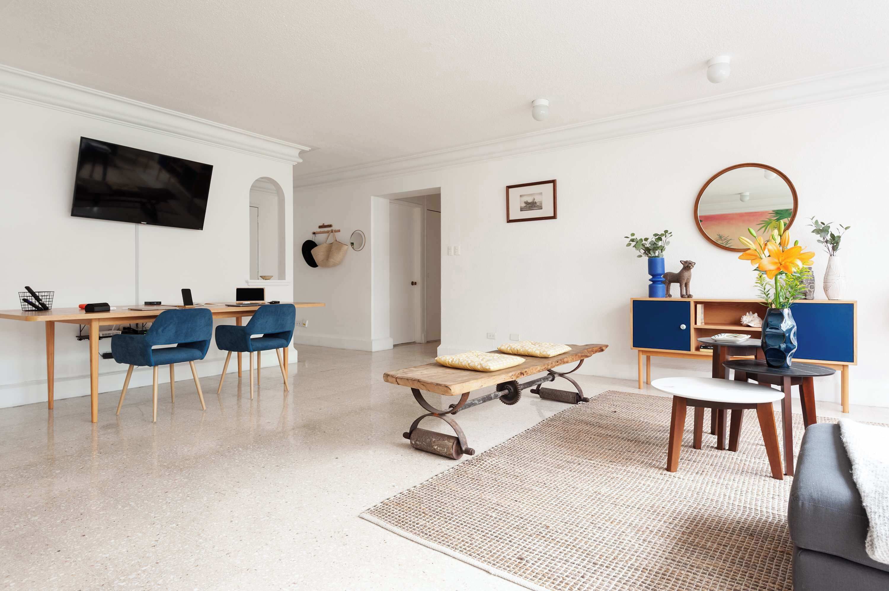 Ein modernes Wohnzimmer mit Flachbildfernseher, einem Essbereich mit Holztisch und blauen Stühlen, einem Couchtisch mit Kissen und einem dekorativen Spiegel.